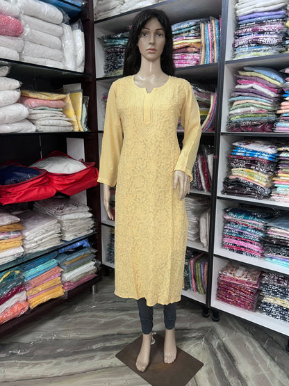 A beige long-sleeve kurta with a textured floral pattern, displayed on a female mannequin in a clothing store. The mannequin is also wearing dark gray leggings, and the background features shelves packed with folded colorful garments.