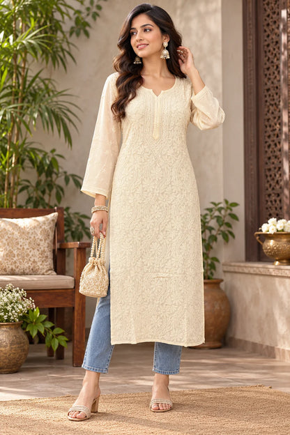 A woman models a cream-colored, long-sleeved kurta with delicate floral embroidery, paired with light blue cropped jeans, holding a small beige handbag in a beautifully decorated indoor-outdoor setting.