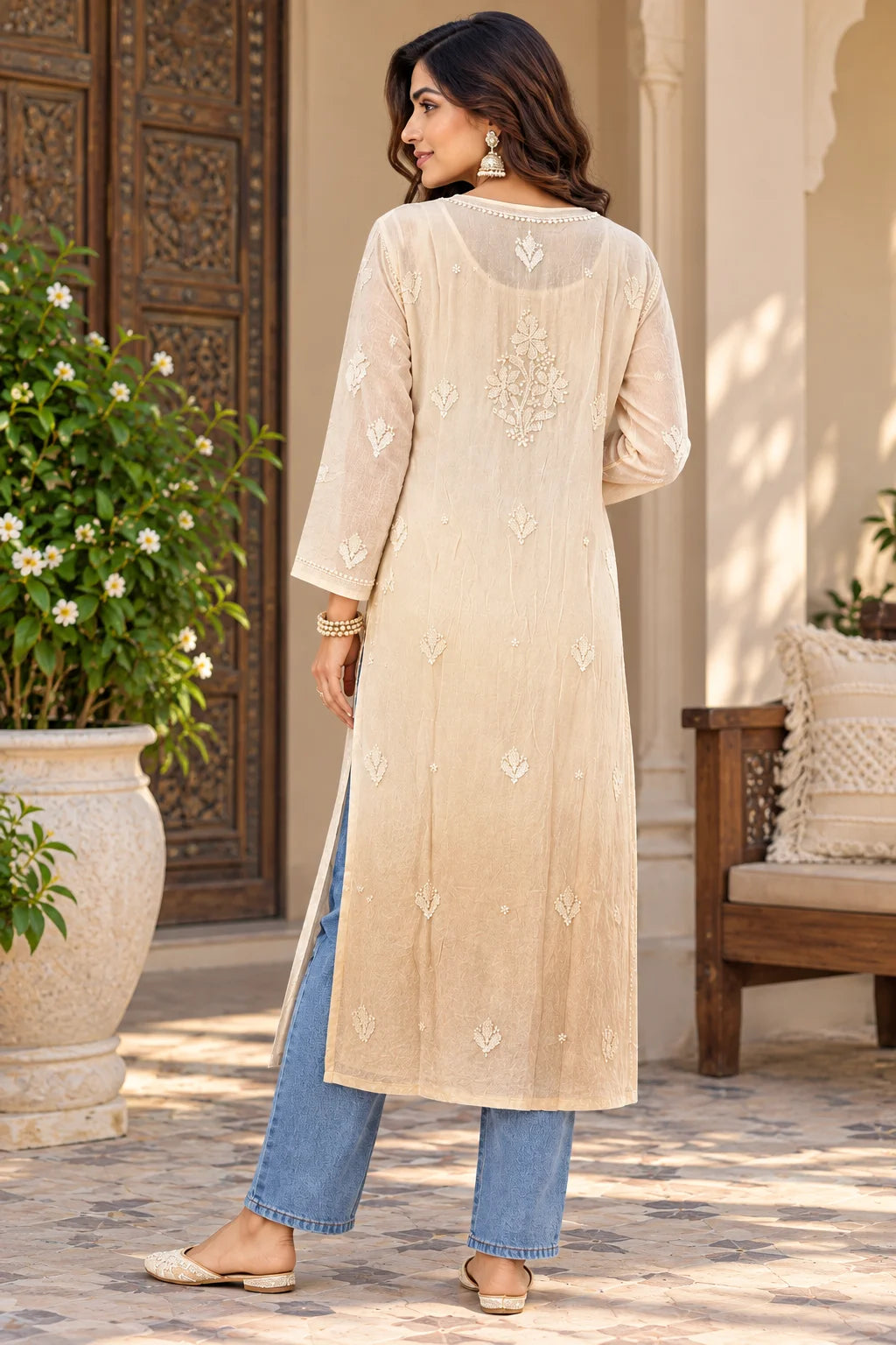 A woman wearing a beige embroidered kurta with blue jeans and flat shoes, photographed from the back in an outdoor courtyard setting.