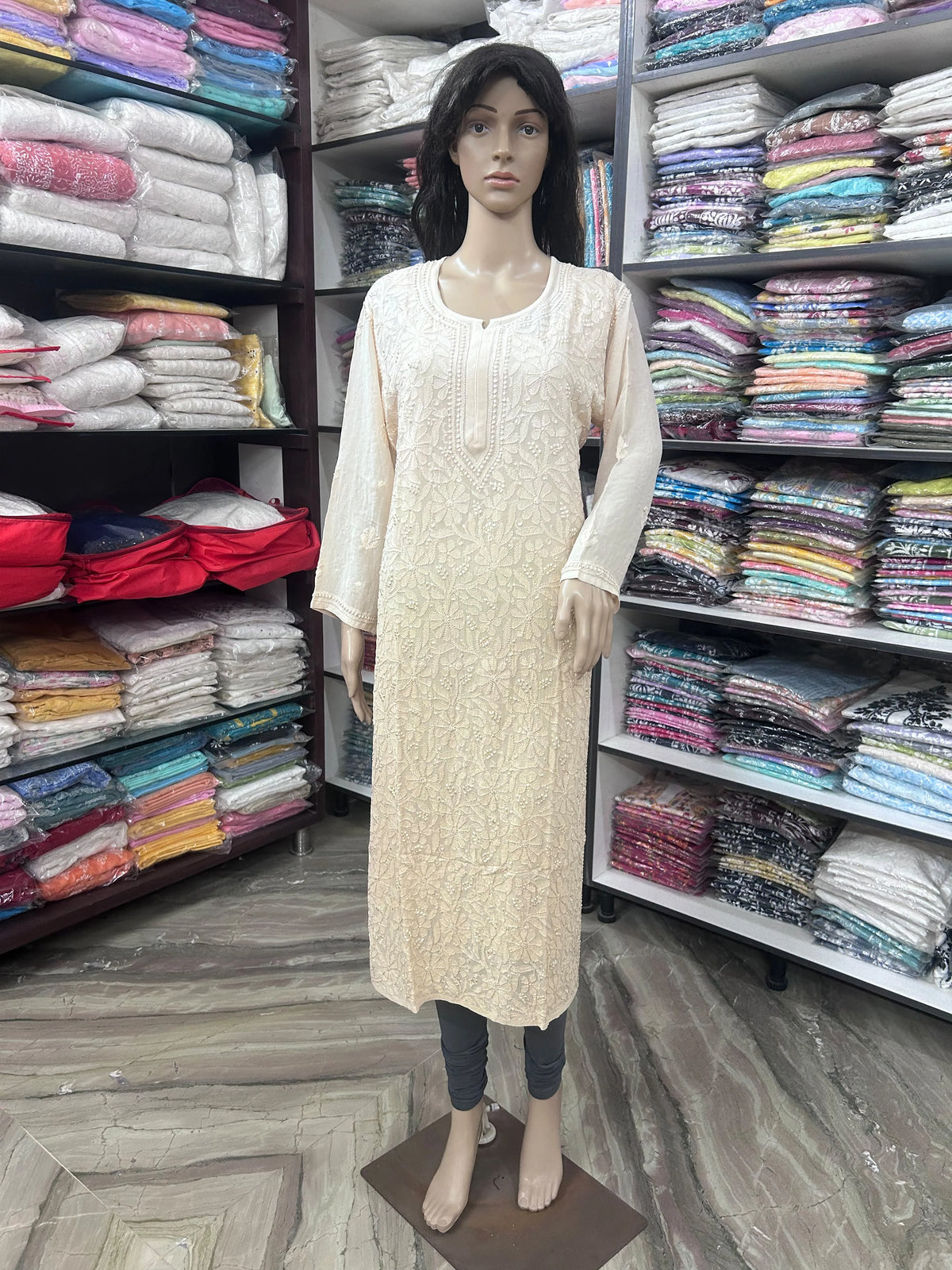 A cream-colored embroidered long kurta displayed on a female mannequin, standing in a retail store with shelves of folded colorful fabrics in the background.