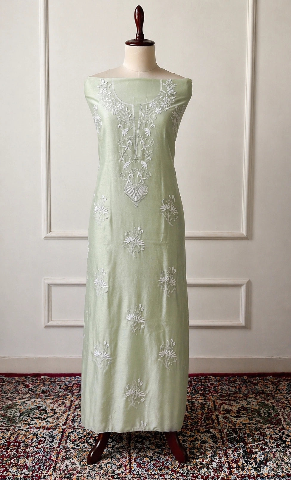 Light green dress with white floral embroidery on a mannequin against a white paneled wall.