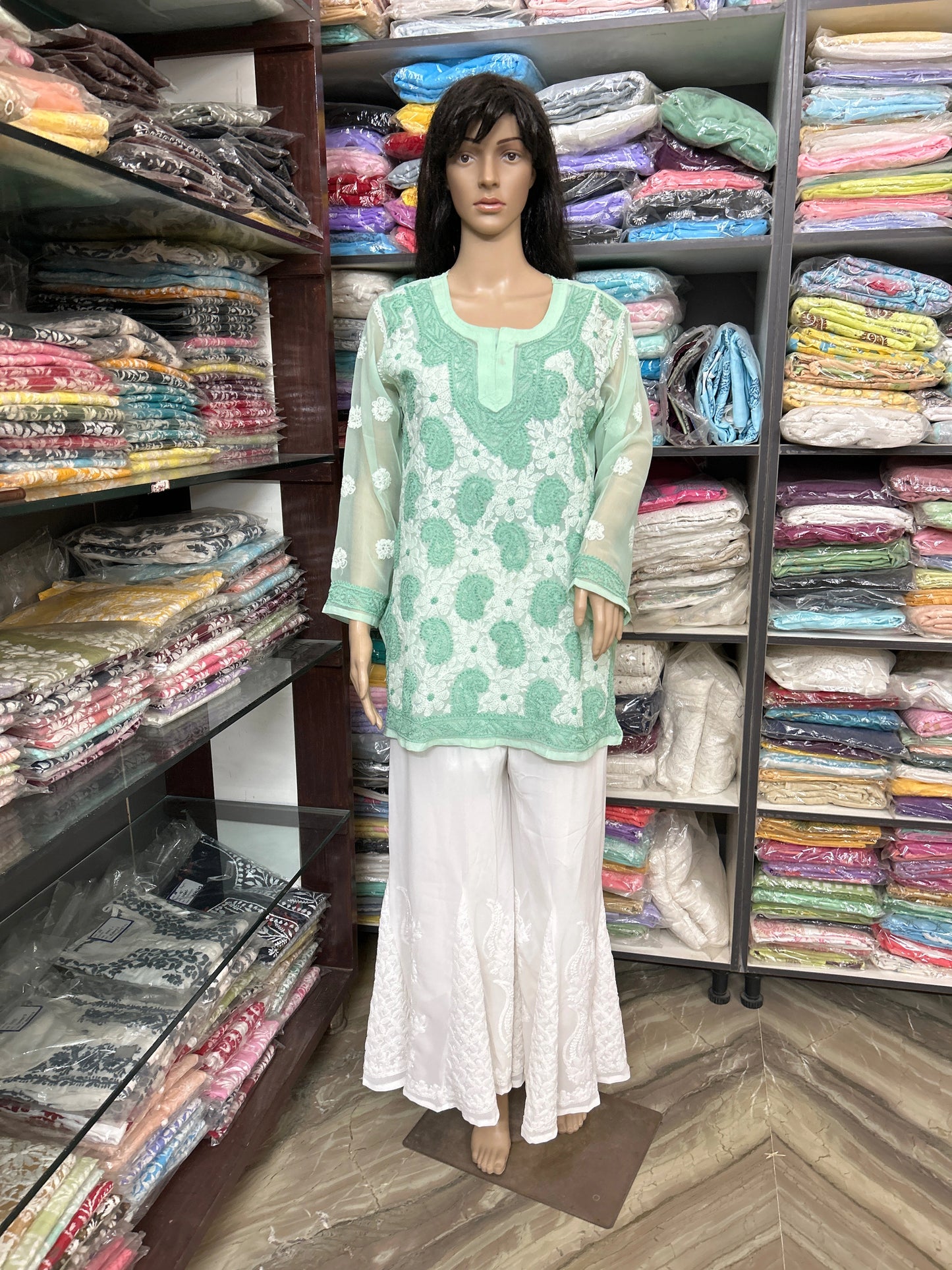 Mannequin wearing a green top and white pants in a fabric store with shelves of colorful fabrics.
