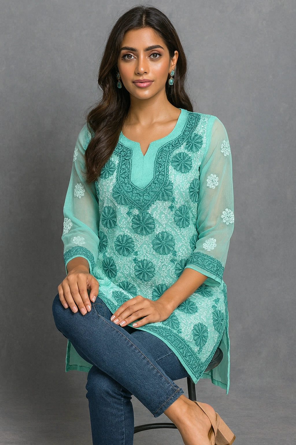 Woman wearing a teal embroidered top sitting on a couch with a vase of pink flowers in the background.