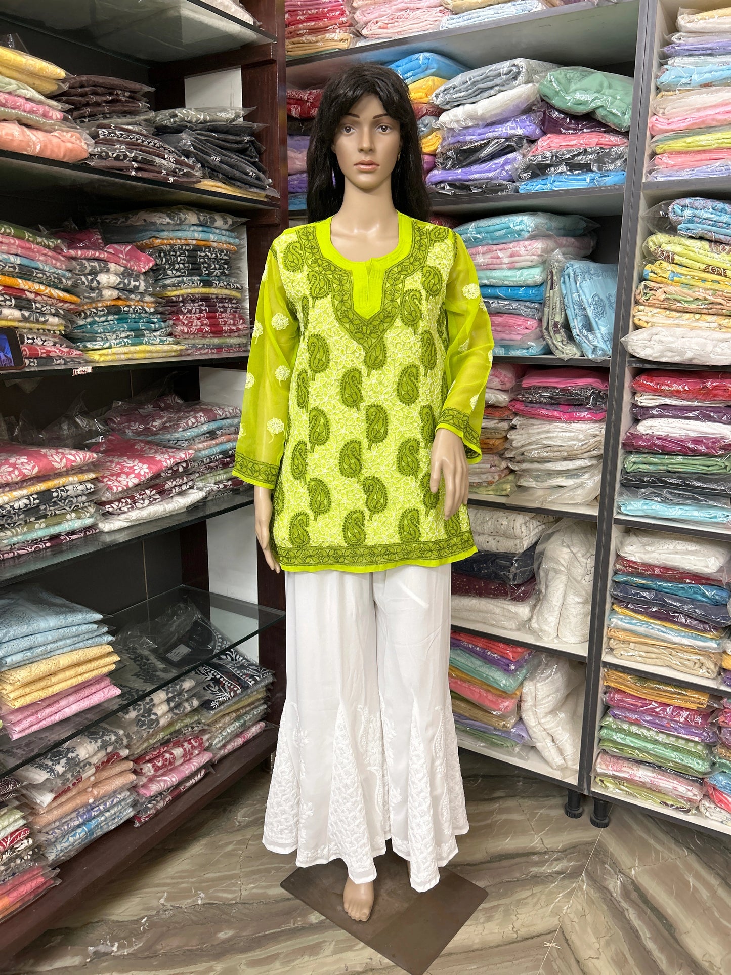 Mannequin wearing a green top and white pants in a fabric store with shelves of colorful fabrics.