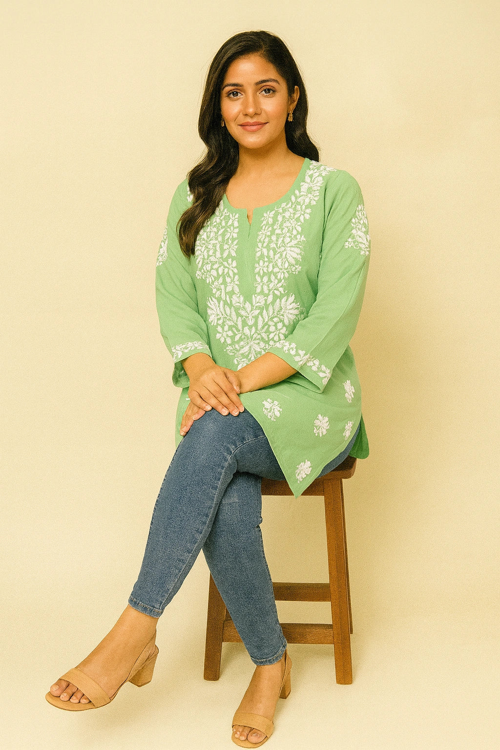Woman wearing a green embroidered top and blue jeans standing against a gray background with a vase of flowers.