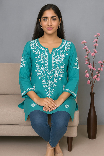 Woman wearing a teal embroidered top sitting on a couch with a vase of pink flowers in the background.