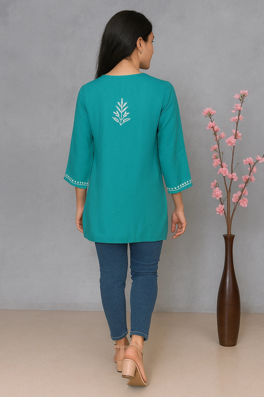 Woman wearing a teal top with floral embroidery, standing against a gray background with a vase of pink flowers.