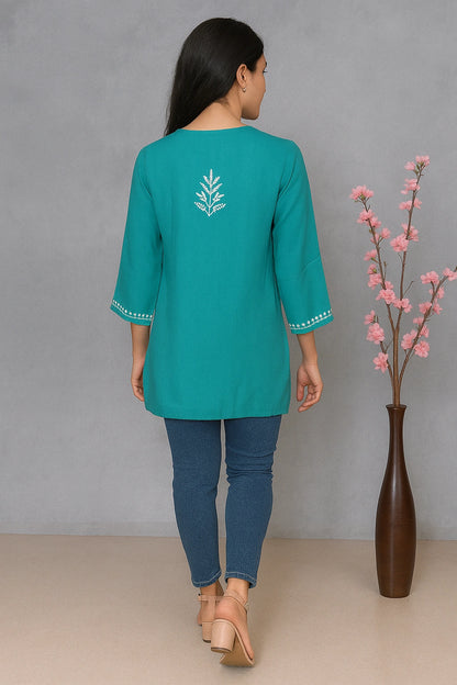 Woman wearing a teal top with floral embroidery, standing against a gray background with a vase of pink flowers.