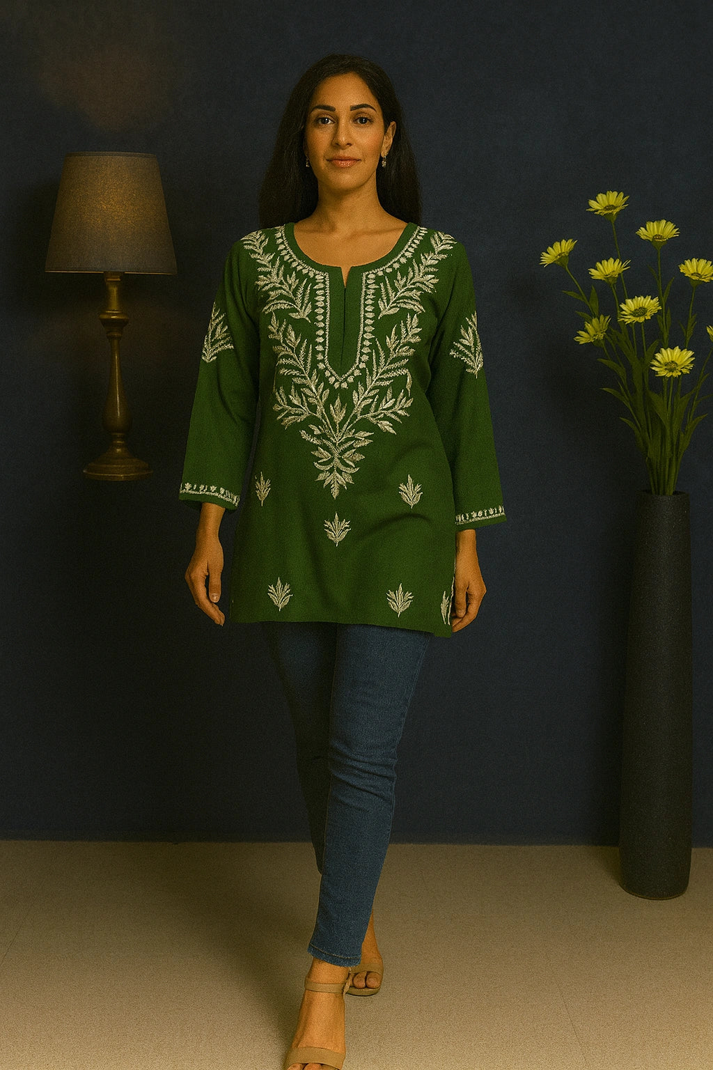 Woman wearing a green embroidered top with blue jeans against a dark wall.