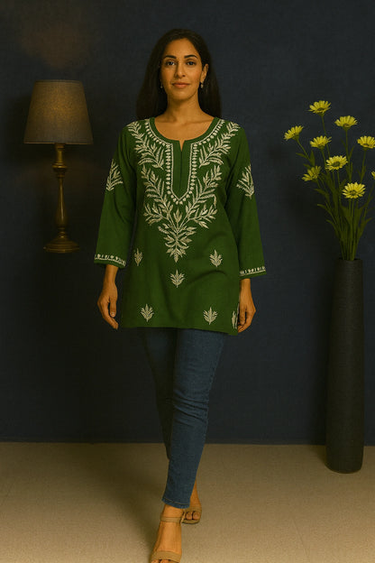 Woman wearing a green embroidered top with blue jeans against a dark wall.