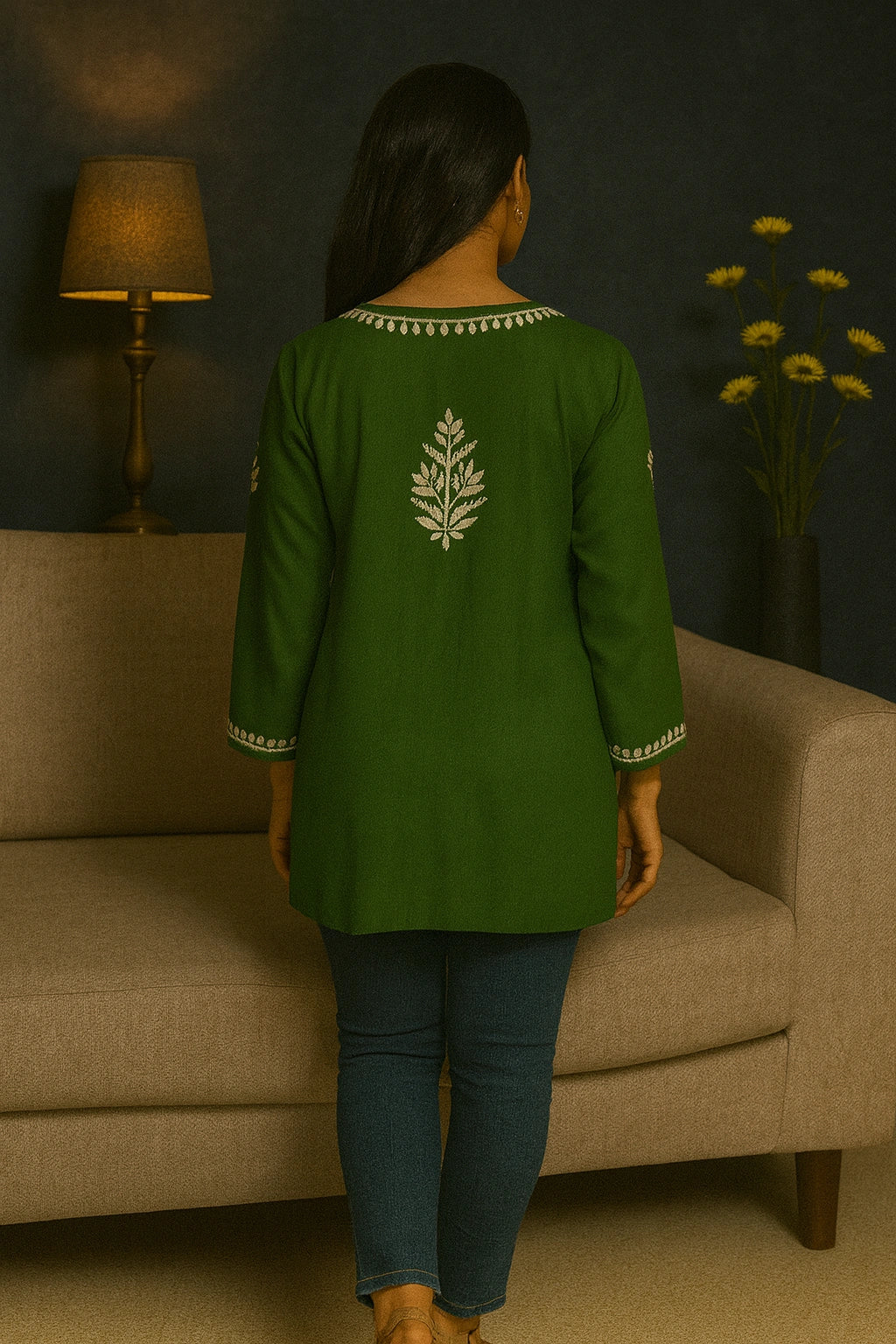 Person wearing a green embroidered top standing in a room with a beige couch and lamp.