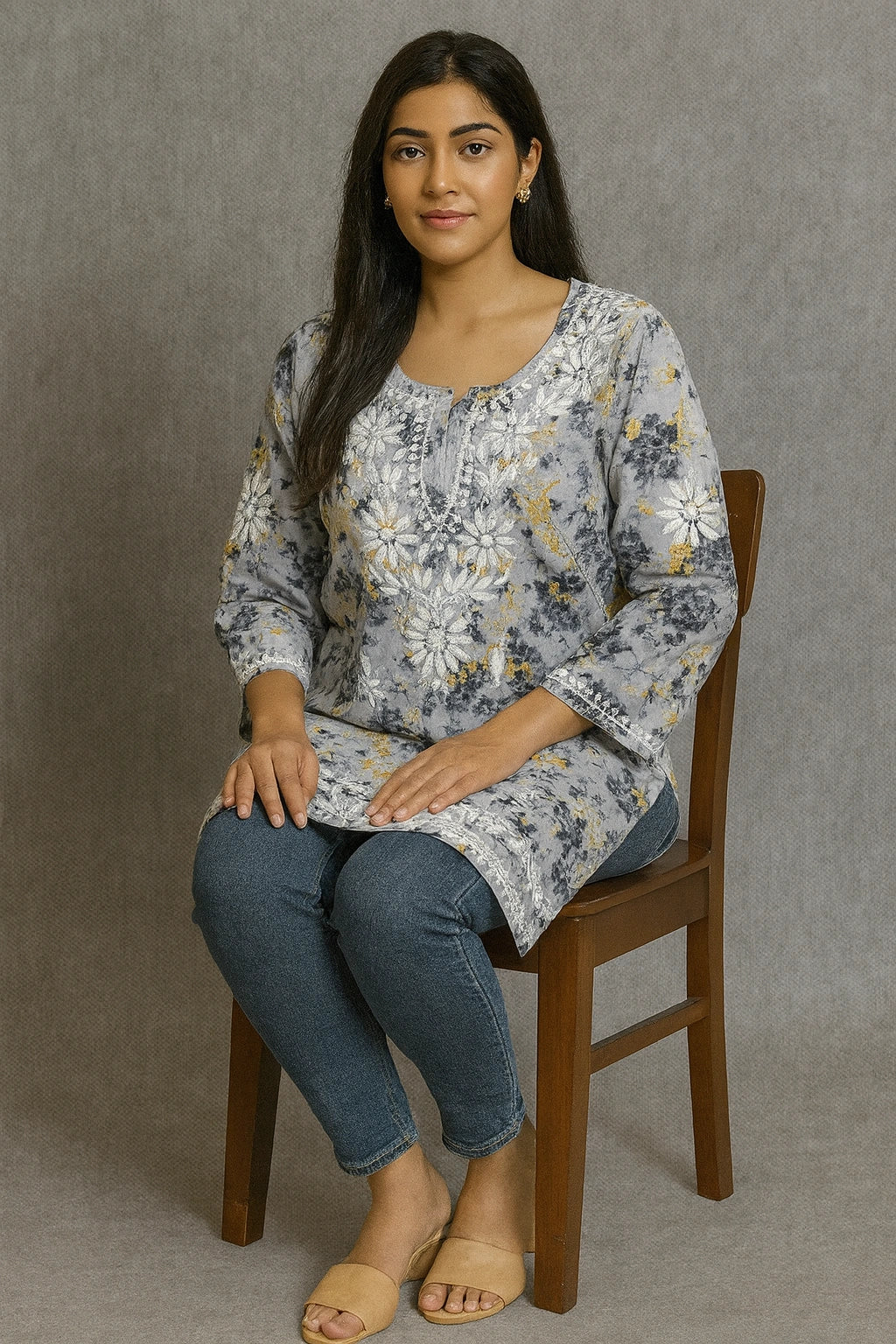 Woman sitting on a lucknowi chikankari wooden chair wearing a floral long-sleeve top and blue jeans against a gray background
