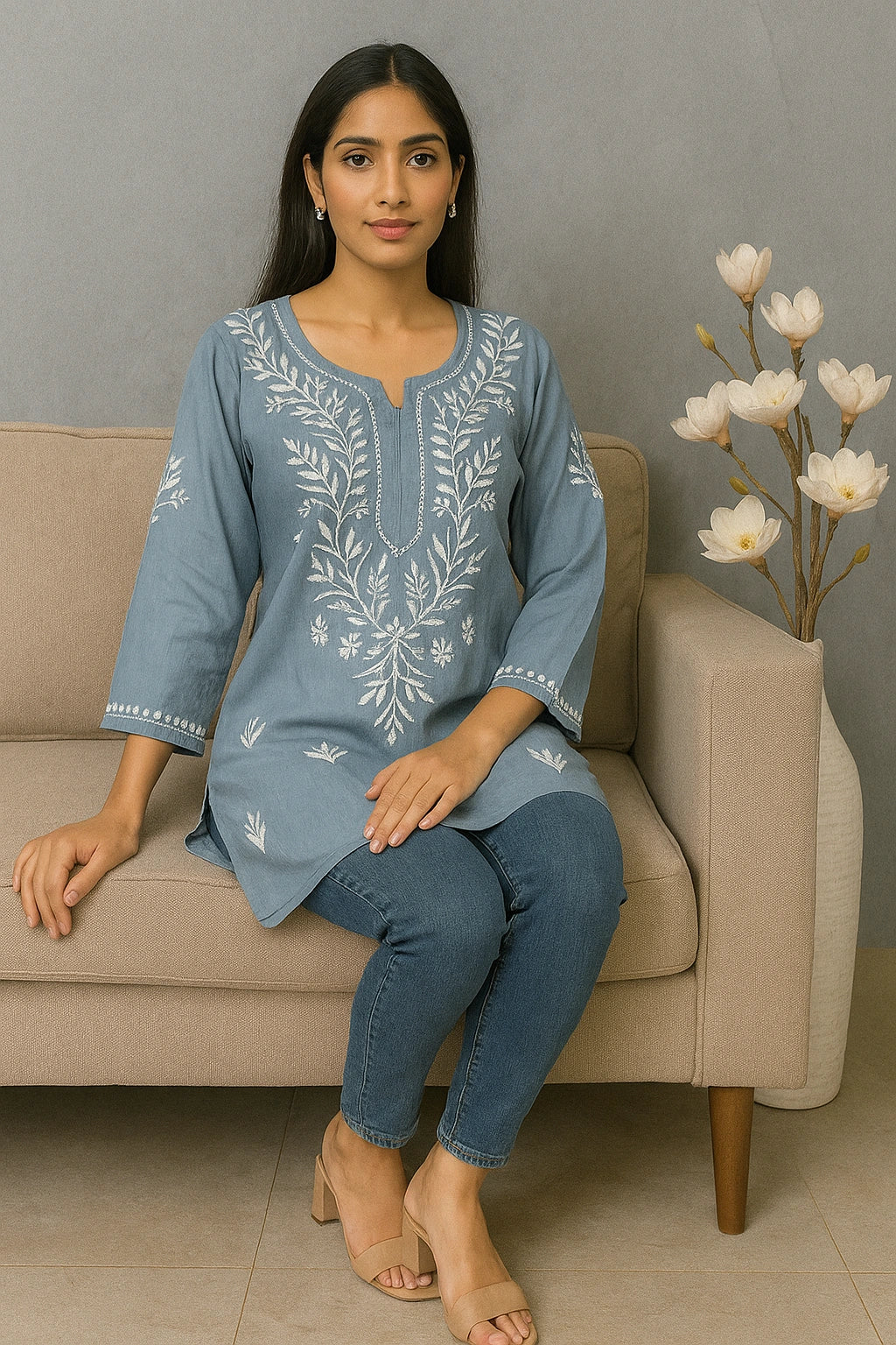 Woman sitting on a beige couch wearing a blue embroidered top and jeans, with a decorative plant in the background.