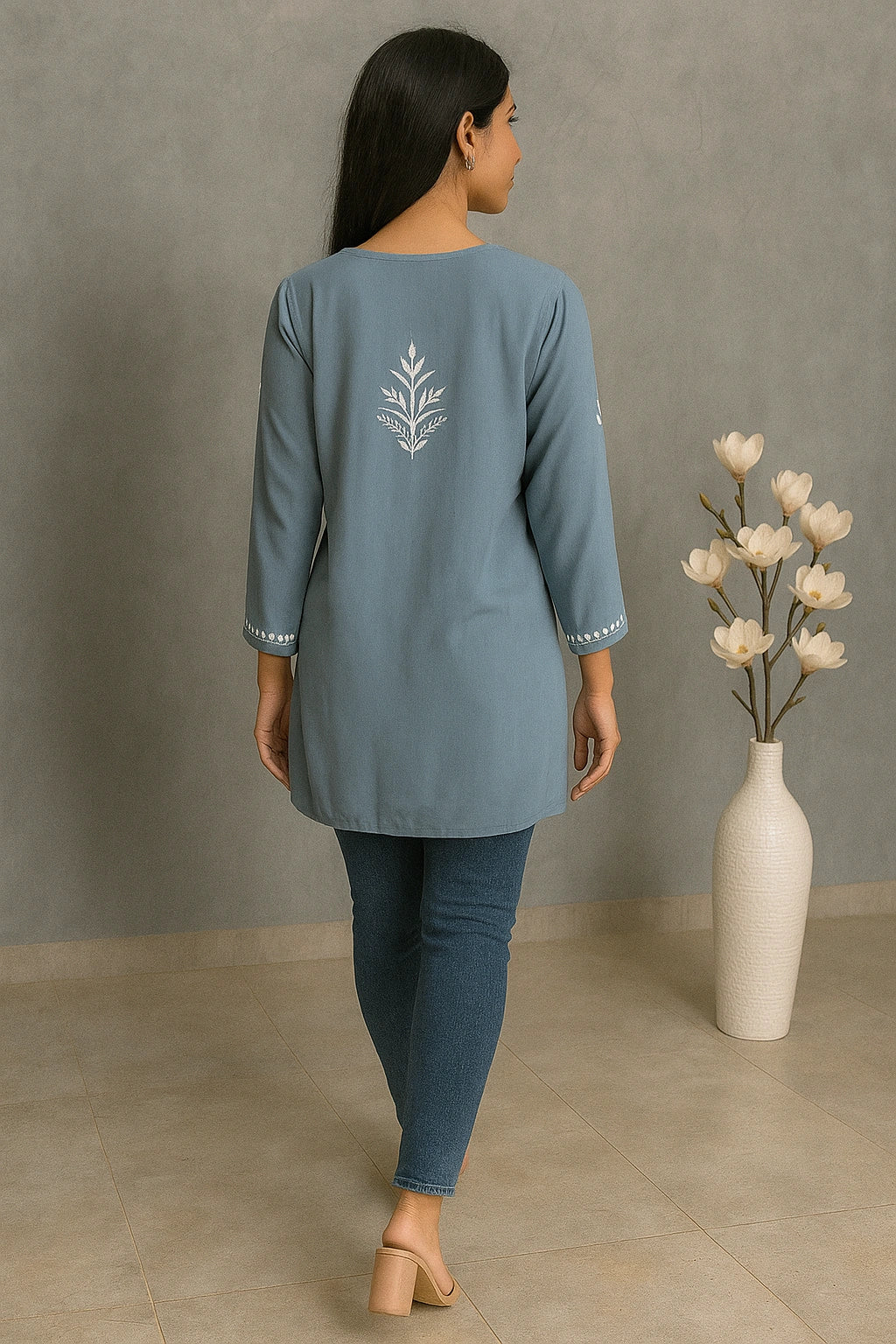Woman wearing a blue outfit with floral embroidery, standing in a room with a vase of flowers.