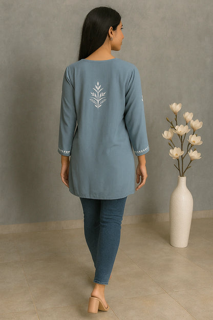 Woman wearing a blue outfit with floral embroidery, standing in a room with a vase of flowers.