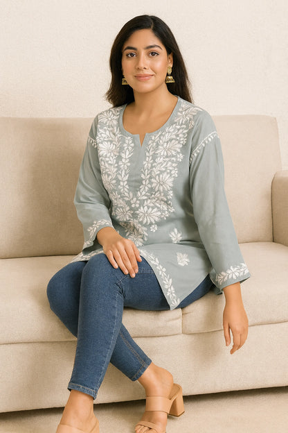 Woman sitting on a beige couch wearing a light blue embroidered top and jeans.