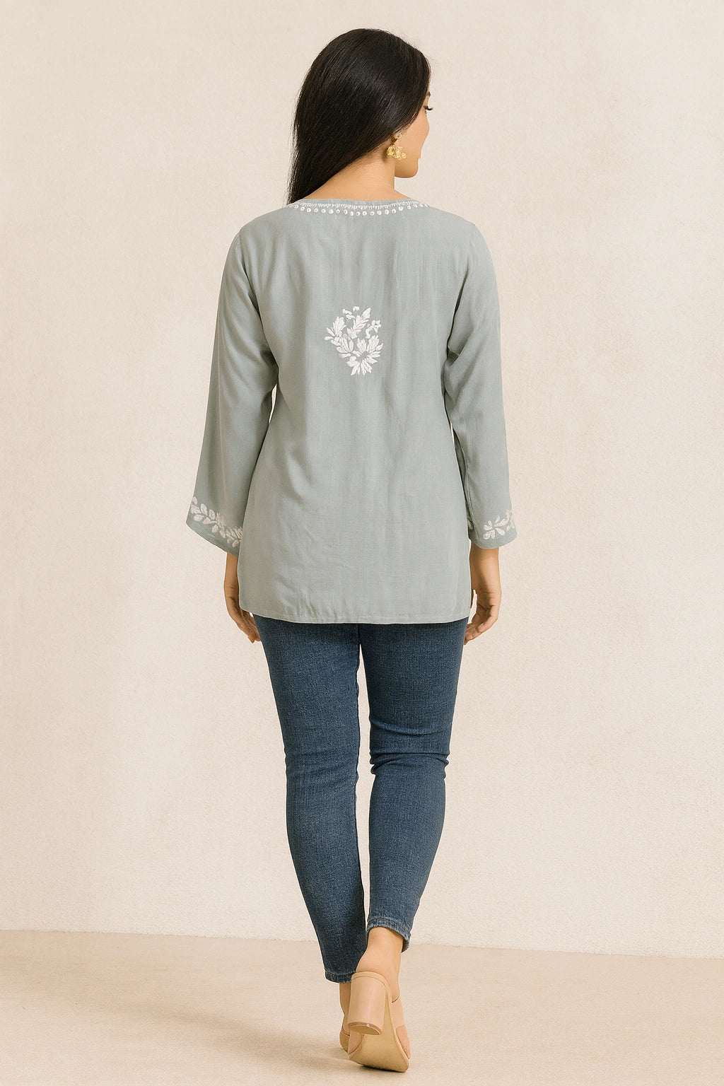 Woman wearing a light gray embroidered top and blue jeans on a beige background