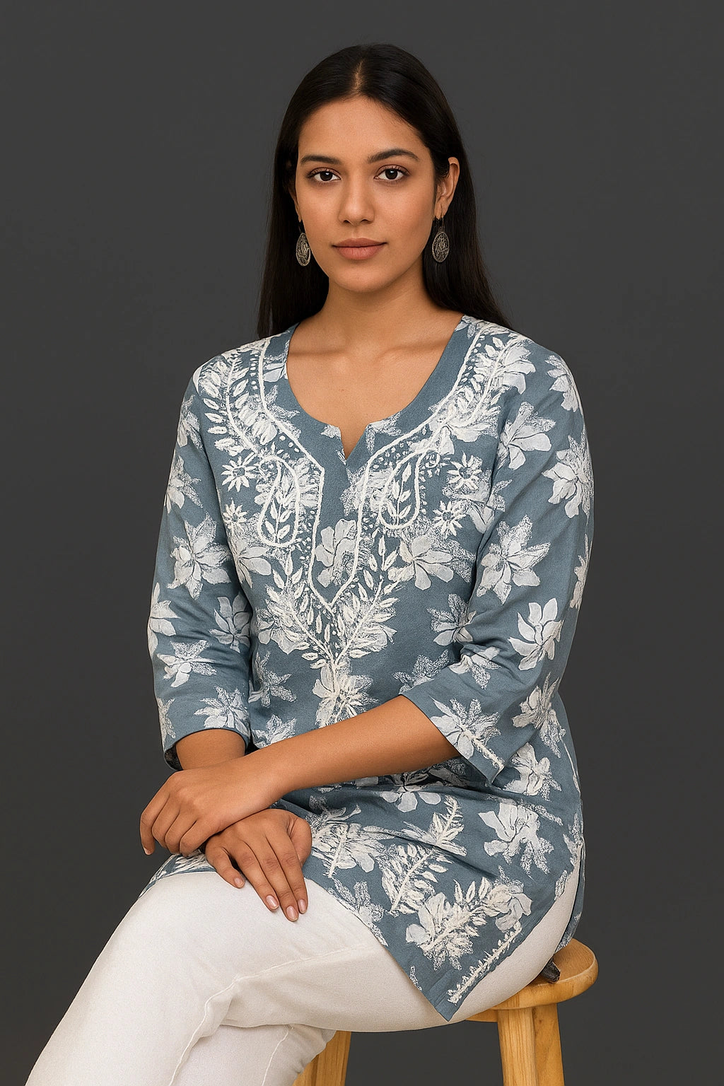 Woman sitting on a chair wearing a grey embroidered top and blue jeans against a gray background