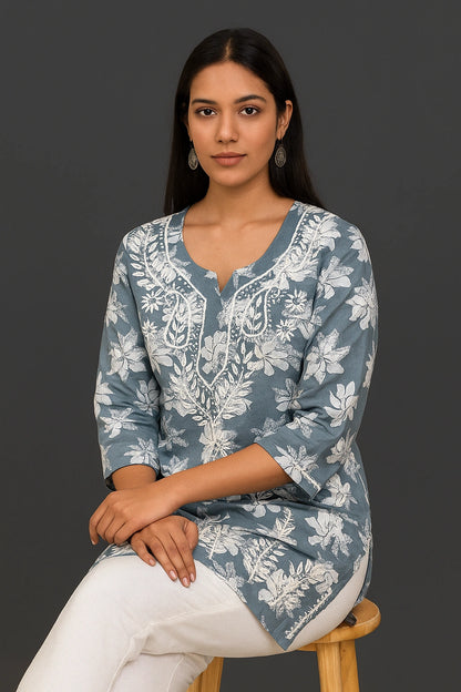 Woman sitting on a chair wearing a grey embroidered top and blue jeans against a gray background