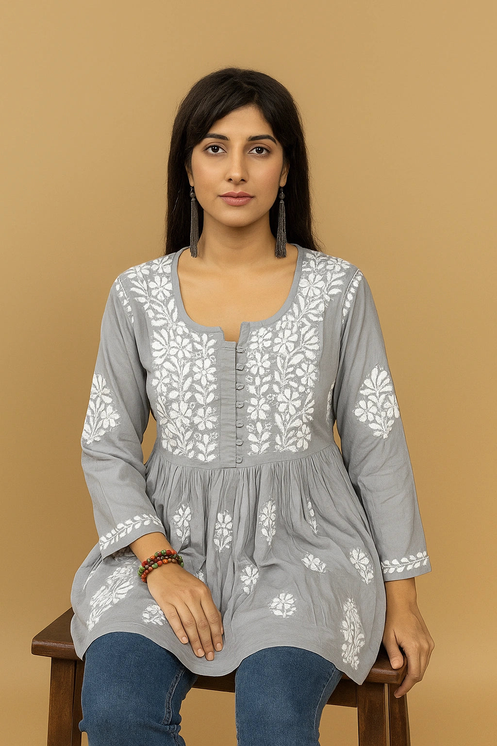 Woman sitting on a chair wearing a grey embroidered top and blue jeans against a gray background