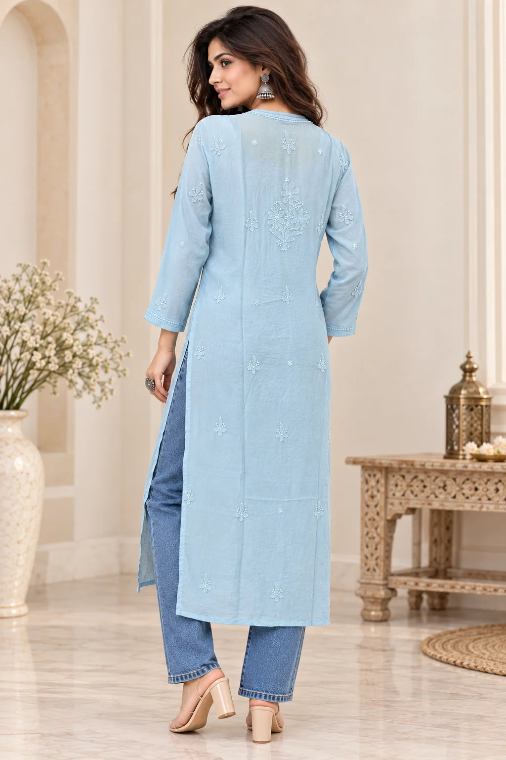 A woman wearing a light blue long kurta with subtle embroidery on the back, paired with blue jeans and beige heels, standing in a decorated indoor setting.