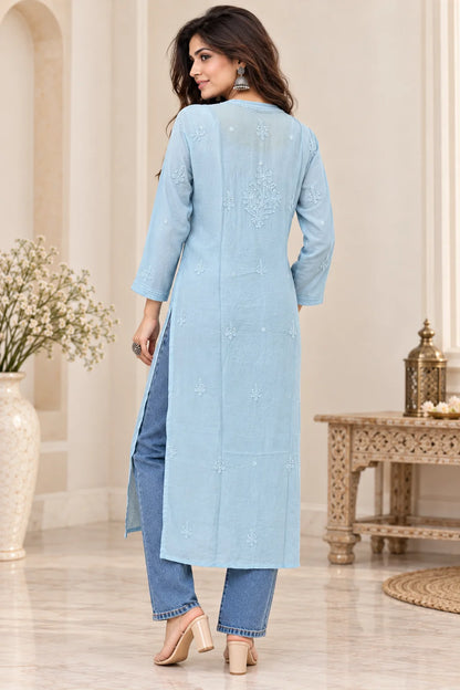 A woman wearing a light blue long kurta with subtle embroidery on the back, paired with blue jeans and beige heels, standing in a decorated indoor setting.