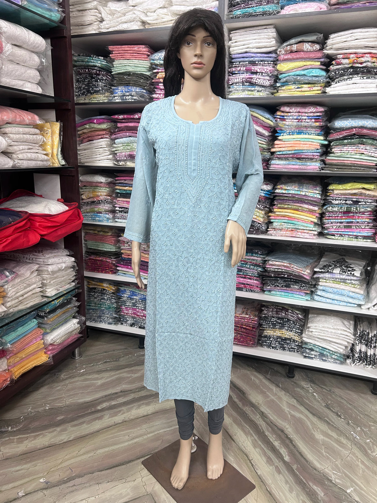 lucknowi chikankari A light blue long kurta with a subtle white floral pattern, displayed on a female mannequin in a clothing store. The mannequin is standing in front of shelves filled with folded garments.