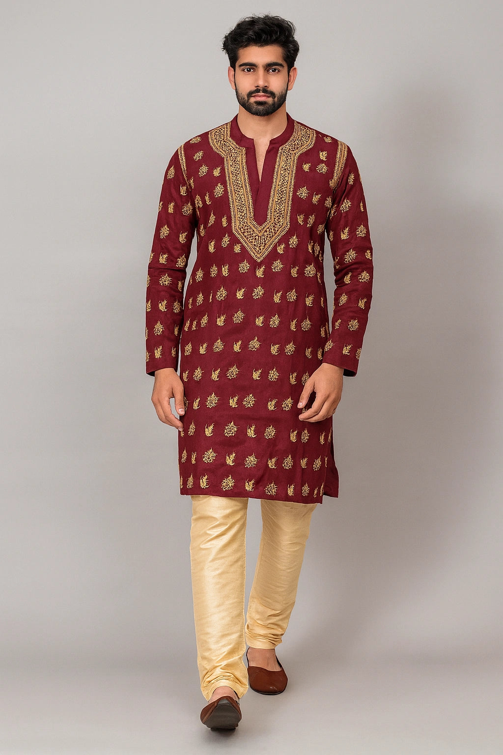 A man modeling a traditional Indian outfit consisting of a maroon kurta with gold embroidery and beige trousers, standing against a neutral gray studio background.