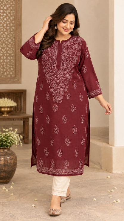 A woman models a three-quarter sleeve maroon kurta with white floral embroidery, paired with off-white trousers and beige flat shoes, in an indoor setting with a beige wall and decorative elements.