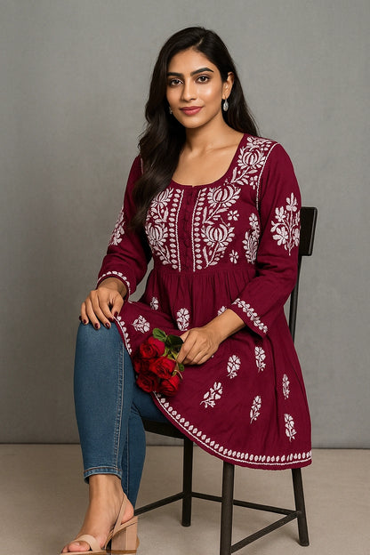 Woman wearing a maroon embroidered dress with blue jeans, sitting on a chair against a gray background.