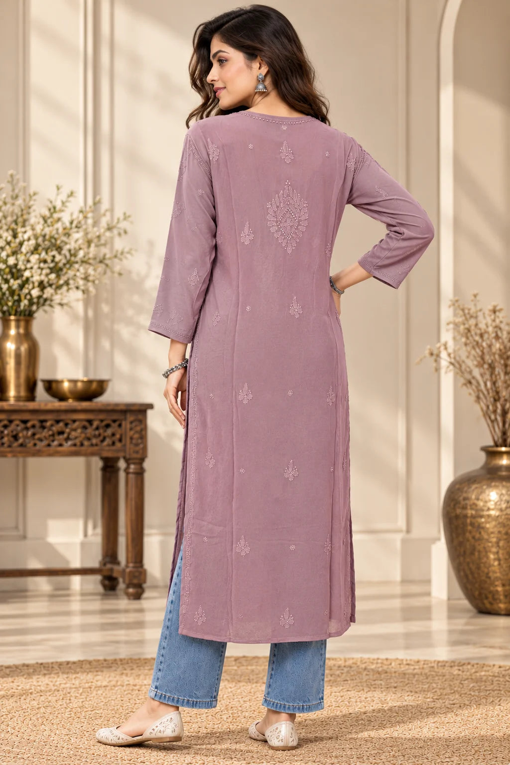 A woman wearing a lavender-purple long kurta with subtle embroidered details, paired with blue jeans and white flat shoes, standing in a decorated indoor setting.