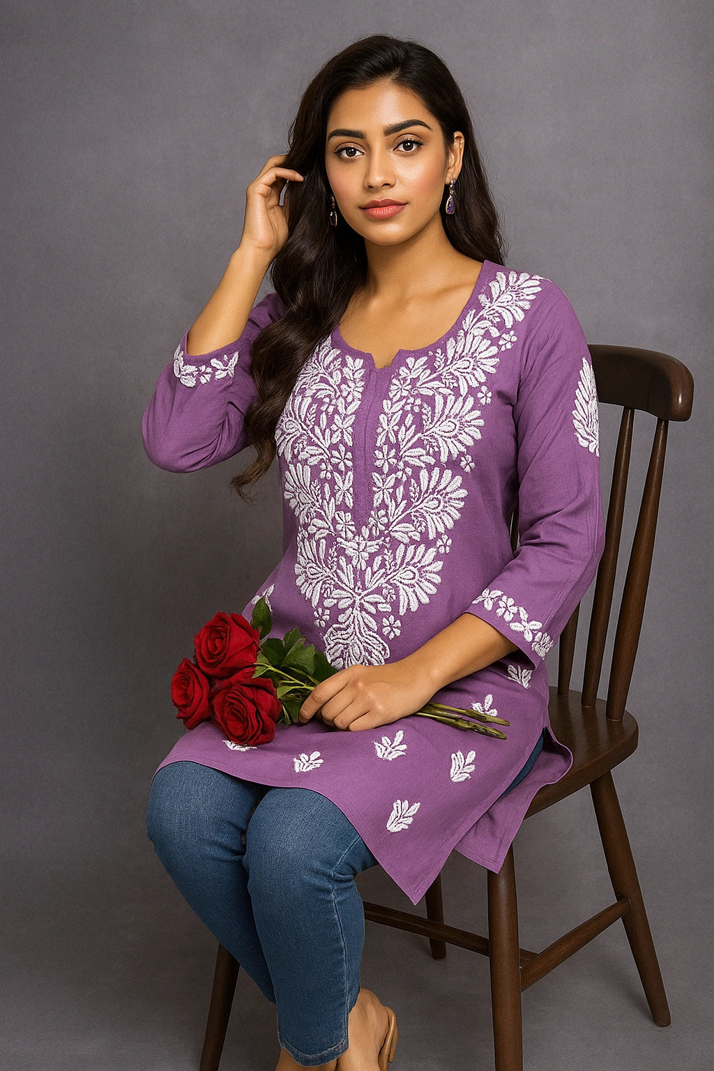 Woman wearing a purple embroidered top sitting on a chair with a gray background