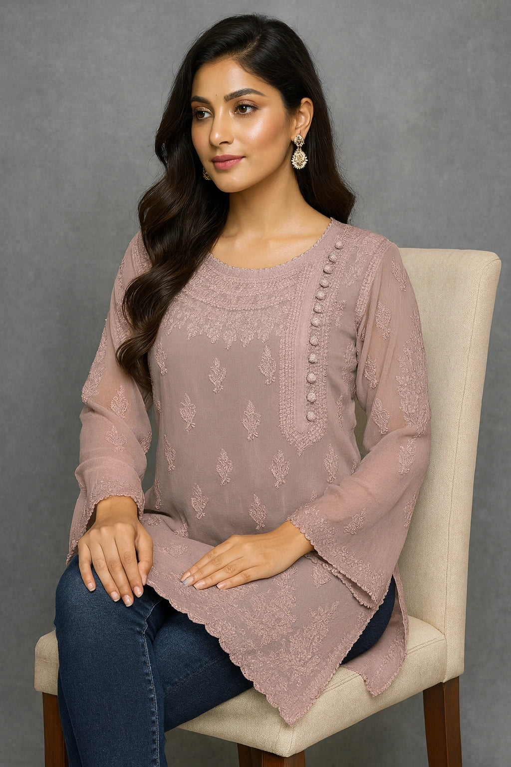 Woman wearing a pink embroidered top sitting on a chair against a gray background