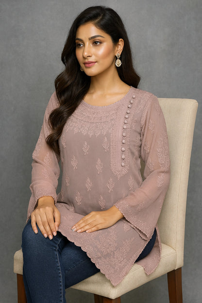 Woman wearing a pink embroidered top sitting on a chair against a gray background