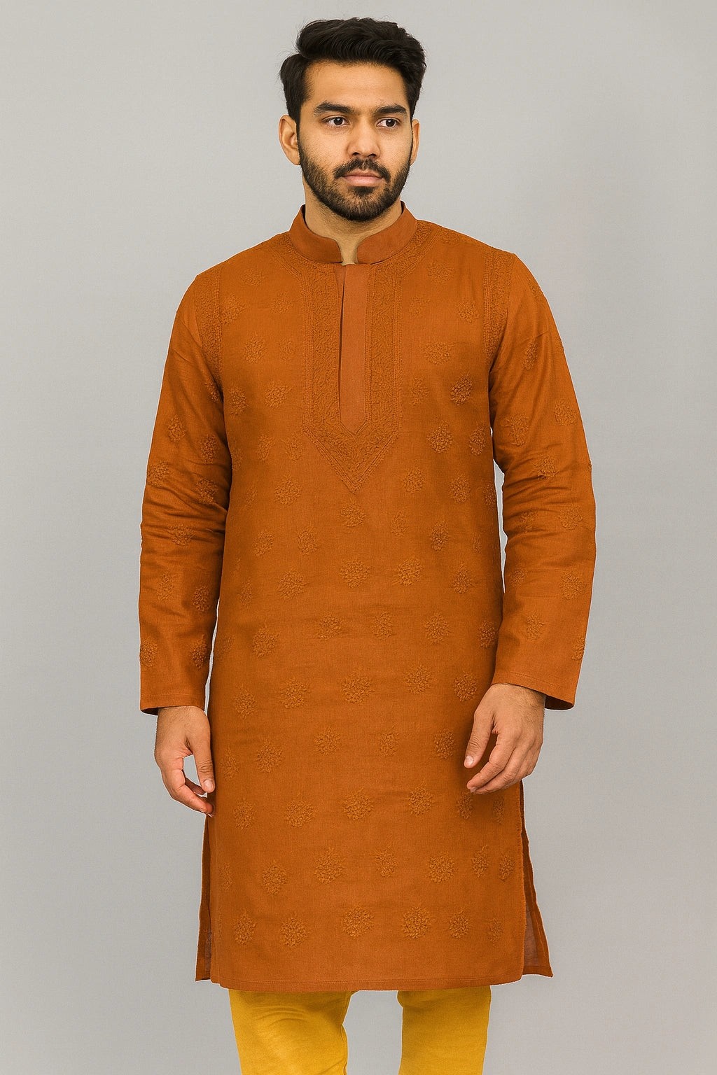A man wearing a rust-orange long-sleeve kurta with subtle tonal embroidery, paired with mustard yellow trousers, standing against a solid gray studio background.