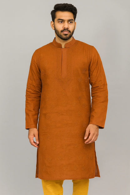 A man wearing a rust-orange long-sleeve kurta with subtle tonal embroidery, paired with mustard yellow trousers, standing against a solid gray studio background.
