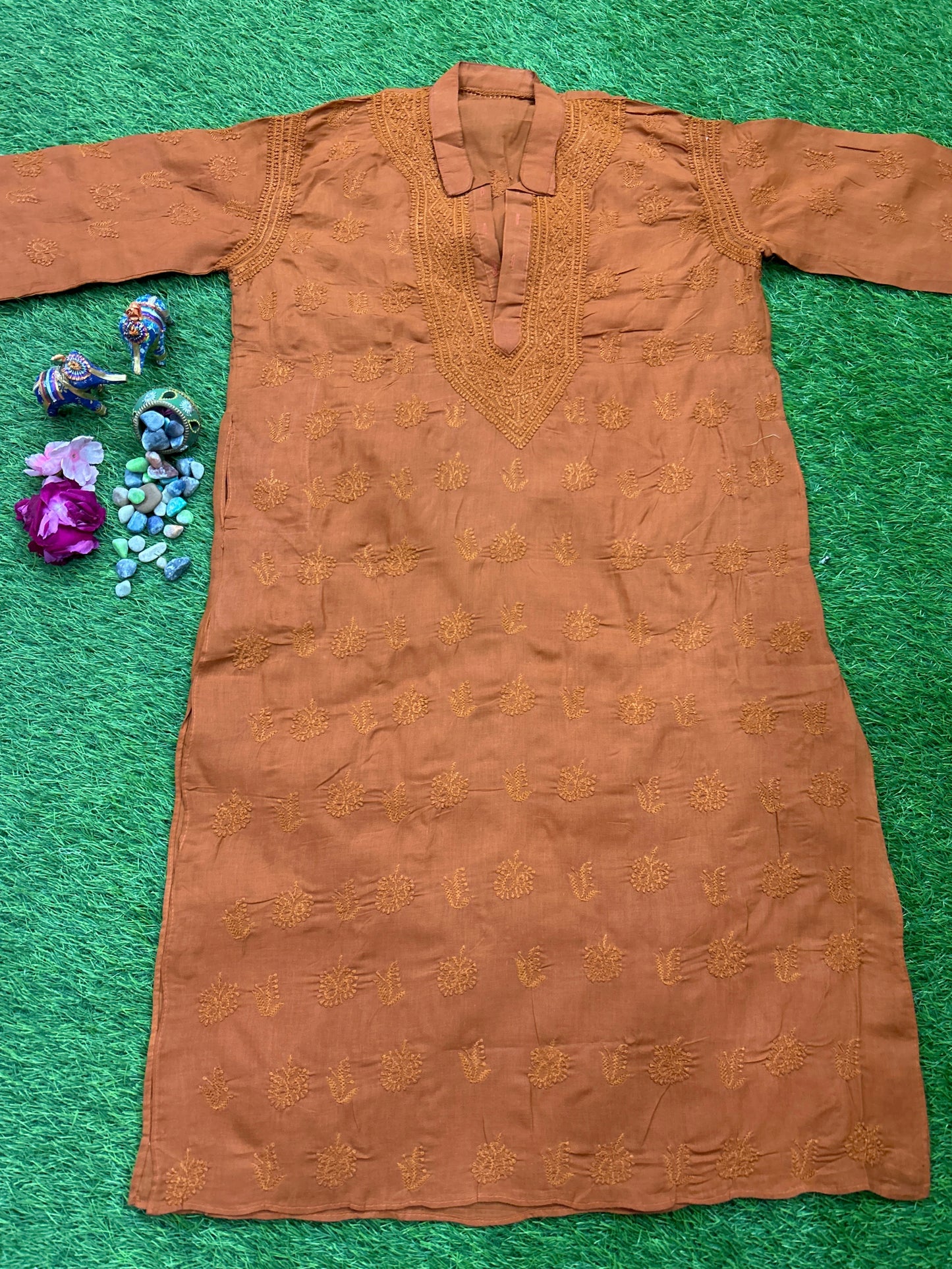 A rust-brown long kurta with gold embroidery, laid flat on a green textured background with small decorative flowers and beads on the left side.