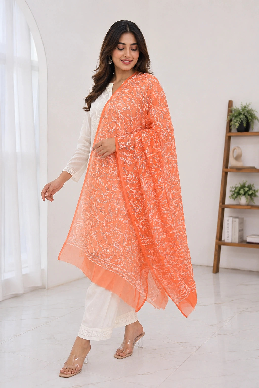 A woman modeling a white traditional outfit with a bright orange embroidered dupatta draped over her shoulder in a bright, modern room.