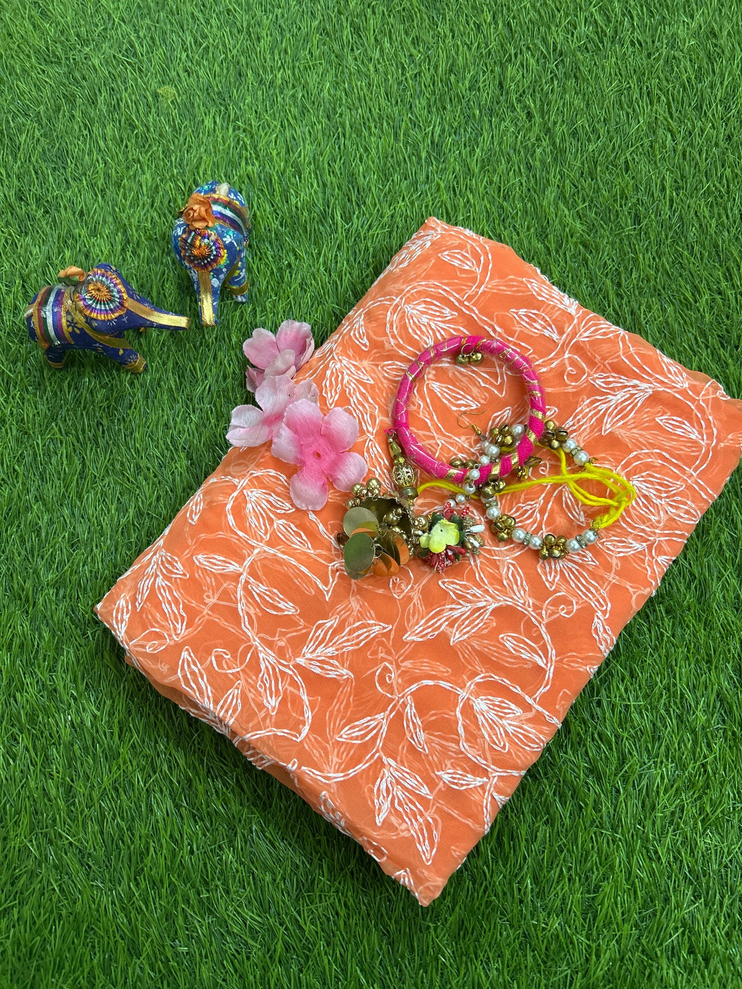 Lucknowi chikankari An orange tepchi work georgette dupatta, styled on green artificial grass with pink flowers, a pink bracelet, and two small decorative elephant figurines.
