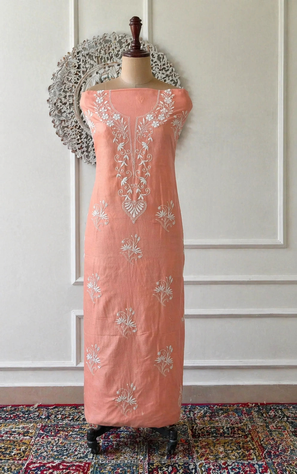 Peach-colored dress with floral embroidery on a mannequin against a white paneled wall.