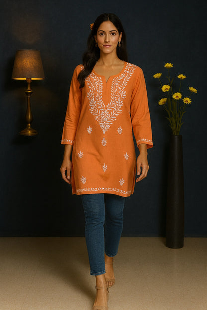 Woman wearing an orange embroidered tunic with blue jeans against a dark wall.