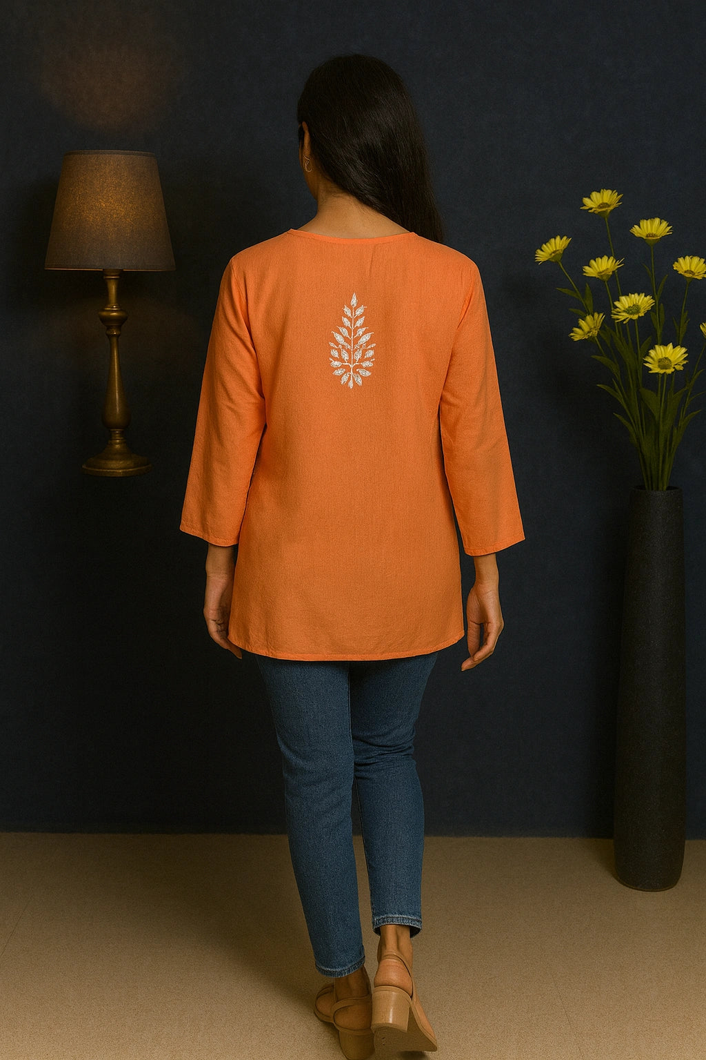 Person wearing an orange top with a leaf design, standing in a room with a dark wall, lamp, and vase of flowers.