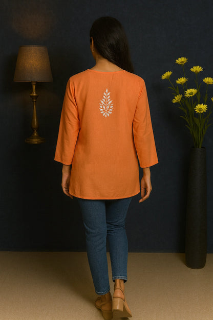 Person wearing an orange top with a leaf design, standing in a room with a dark wall, lamp, and vase of flowers.