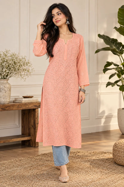 A woman models a light peach embroidered kurta with three-quarter sleeves and a round neckline, paired with blue jeans and beige heels in a bright, modern indoor setting.