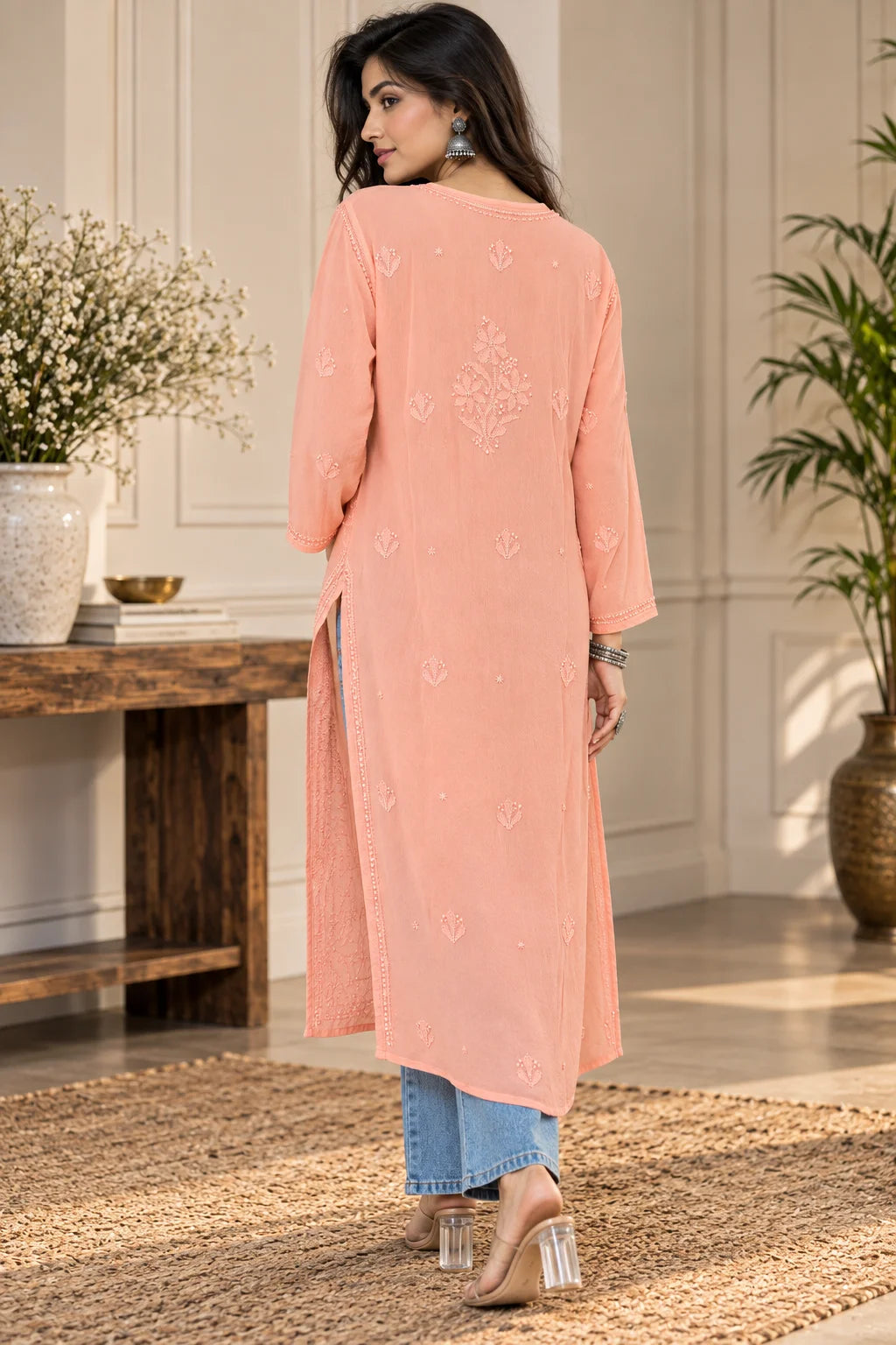 A woman wearing a light peach long kurta with subtle embroidery on the back, paired with blue jeans and beige heels, standing in a decorated indoor setting.