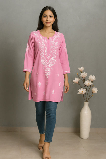 Woman wearing a pink embroidered kurta with blue jeans standing next to a vase with flowers against a gray background.