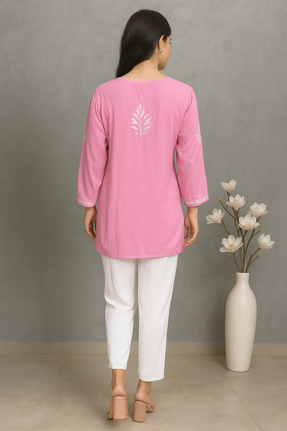 Woman wearing a lucknowi chikankari pink top with a white floral design, standing next to a vase with flowers against a gray wall.