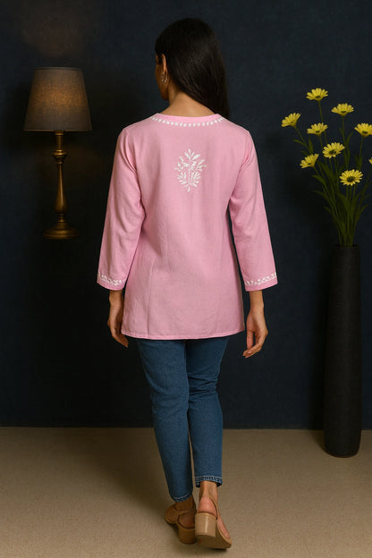 Person wearing a pink long-sleeve top with floral embroidery, standing against a dark wall with a lamp and flowers.