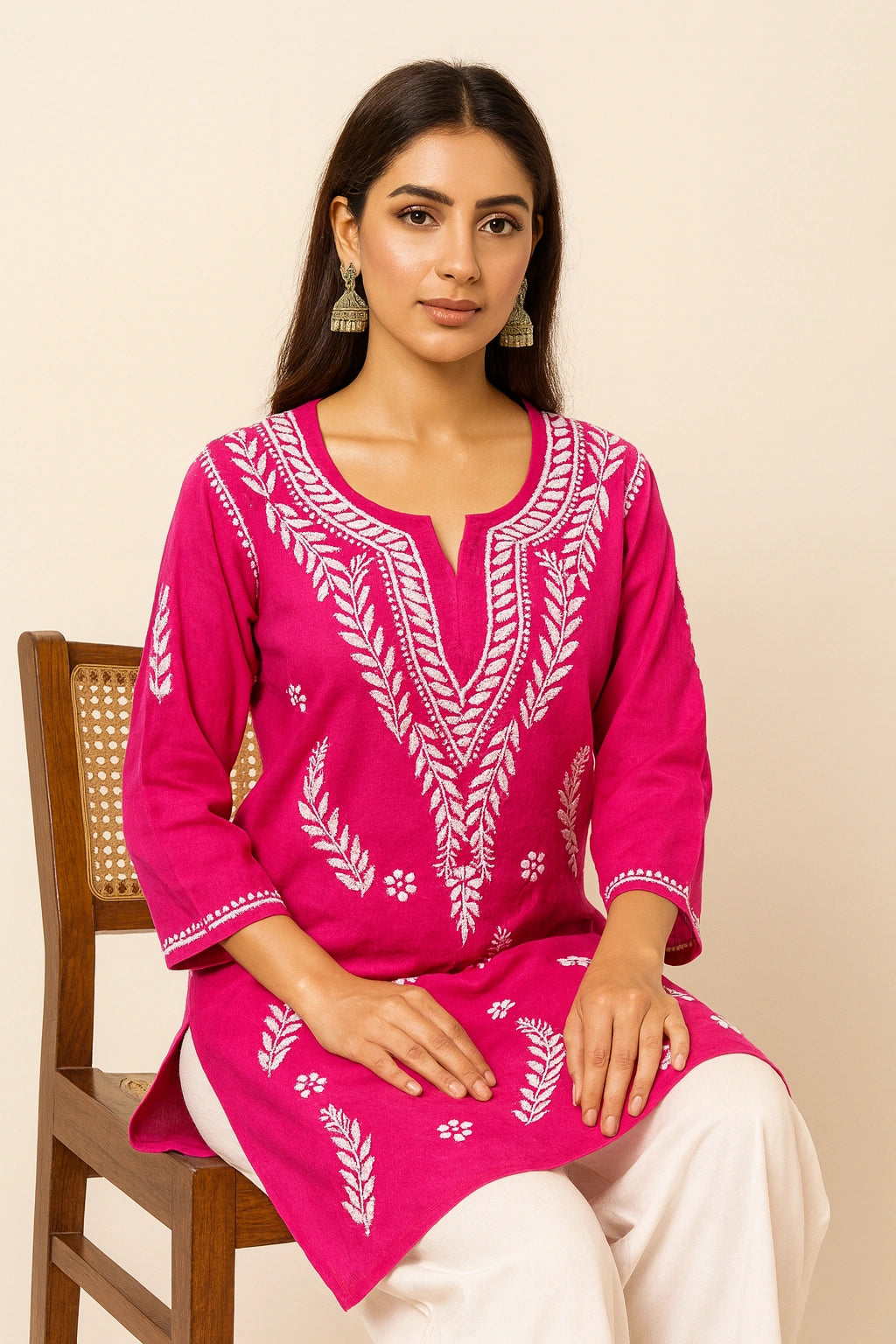 Woman wearing a pink embroidered top sitting on a wooden chair against a beige background