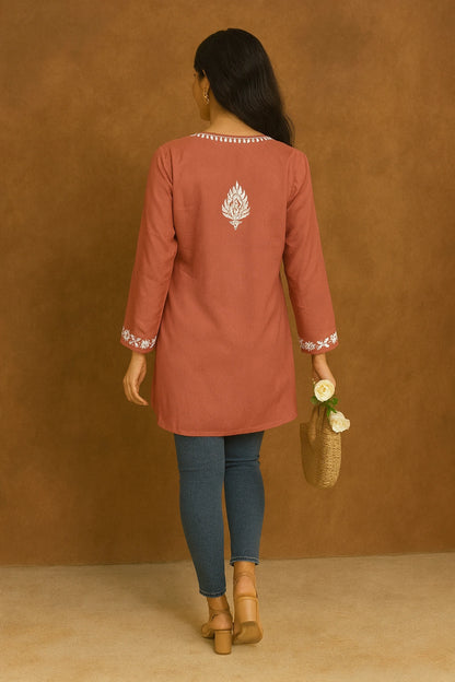 Woman wearing a rust-colored embroidered top with blue pants against a brown background